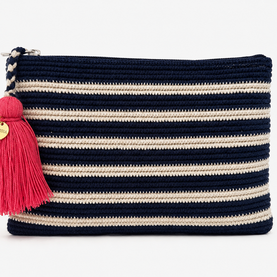 A crochet zip pouch with horizontal navy blue and cream stripes, featuring a bright pink tassel charm with a gold-tone circular brand tag, set against a plain white background.
