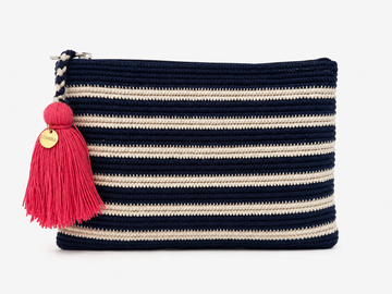 A crochet zip pouch with horizontal navy blue and cream stripes, featuring a bright pink tassel charm with a gold-tone circular brand tag, set against a plain white background.