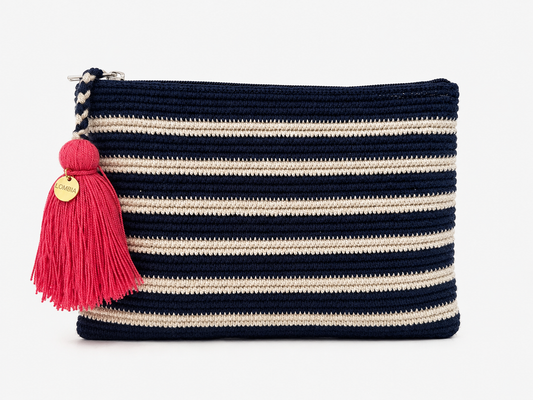 A crochet zip pouch with horizontal navy blue and cream stripes, featuring a bright pink tassel charm with a gold-tone circular brand tag, set against a plain white background.