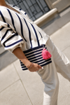 A person outdoors wearing a cream knit cardigan with navy blue stripes, cream trousers, and carrying a matching cream and navy striped woven clutch with a bright pink tassel.