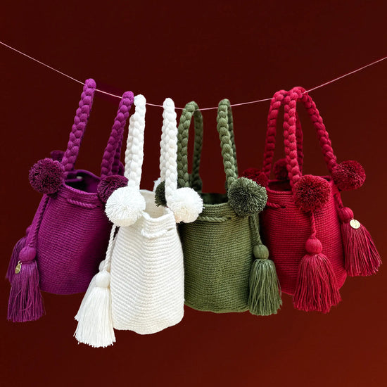Four colorful, Christmas-perfect crochet bags with pom-poms and tassels hanging on a string against a red background, made in Colombia by Wayuu artisans