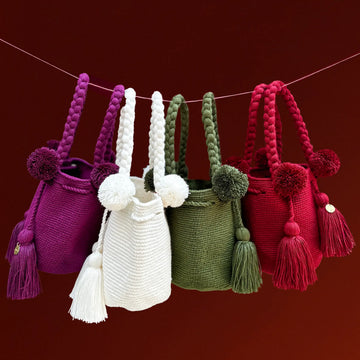 Four colorful crochet bags with pom-poms and tassels hanging on a string against a red background, made in Colombia by Wayuu artisans