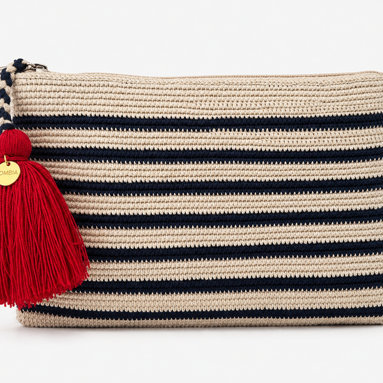 A woven rectangular crochet clutch bag in beige with horizontal stripes in navy blue, featuring a large red tassel with a gold round charm on the zipper pull.