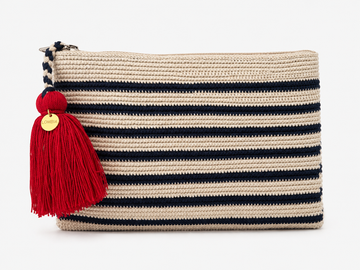 A woven rectangular crochet clutch bag in beige with horizontal stripes in navy blue, featuring a large red tassel with a gold round charm on the zipper pull.