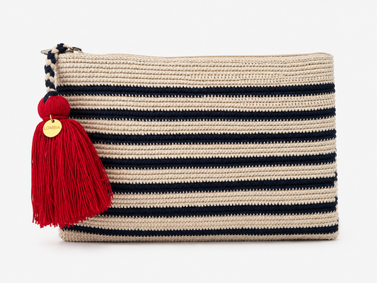 A woven rectangular crochet clutch bag in beige with horizontal stripes in navy blue, featuring a large red tassel with a gold round charm on the zipper pull.